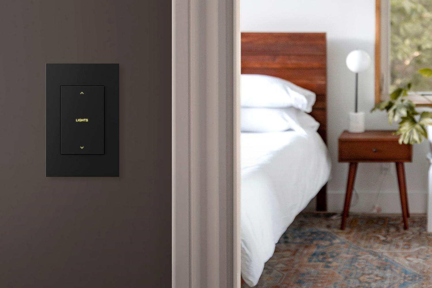 A close-up of a sleek, modern light switch on a dark wall, with a bedroom visible in the background featuring a wooden bed and a side table with a lamp.