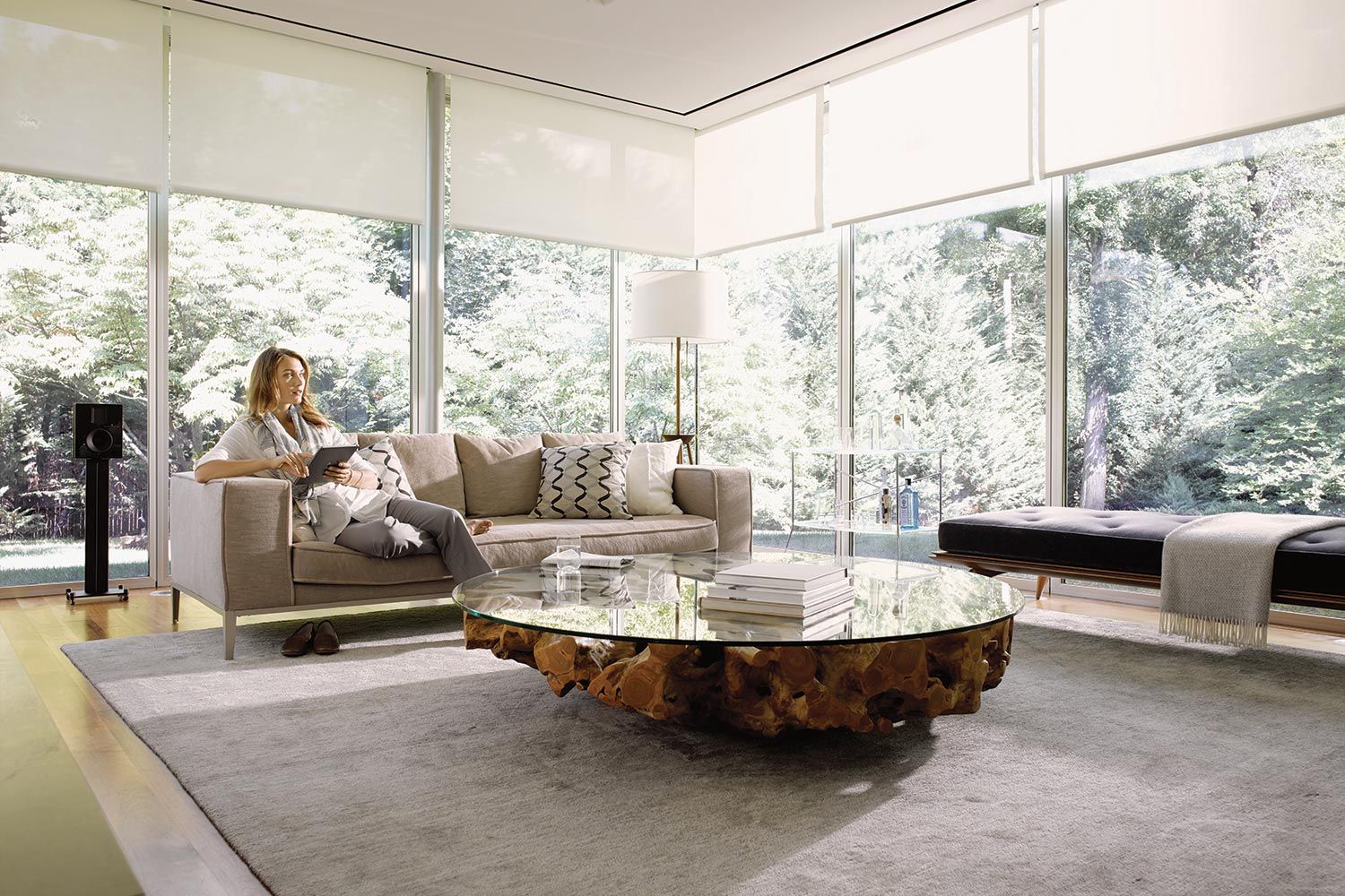 A woman sits on a beige couch in a modern living room with floor-to-ceiling windows, holding a tablet and relaxing. The room features a unique glass coffee table with a wooden base and a light grey rug.
