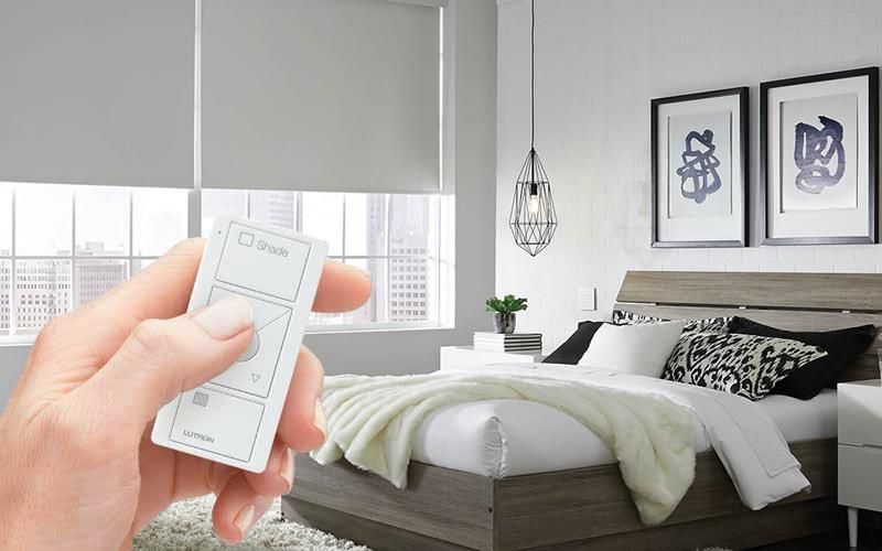 A hand holding a Lutron remote control in a modern bedroom with roller shades, highlighting single-room control capabilities.