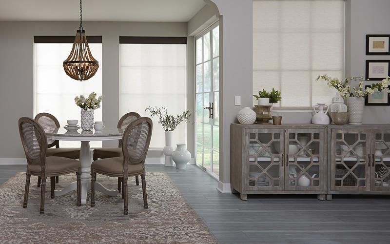 A dining room with Lutron roller shades on large windows, elegantly furnished with a round table, chairs, and a chandelier, illustrating energy-saving features.