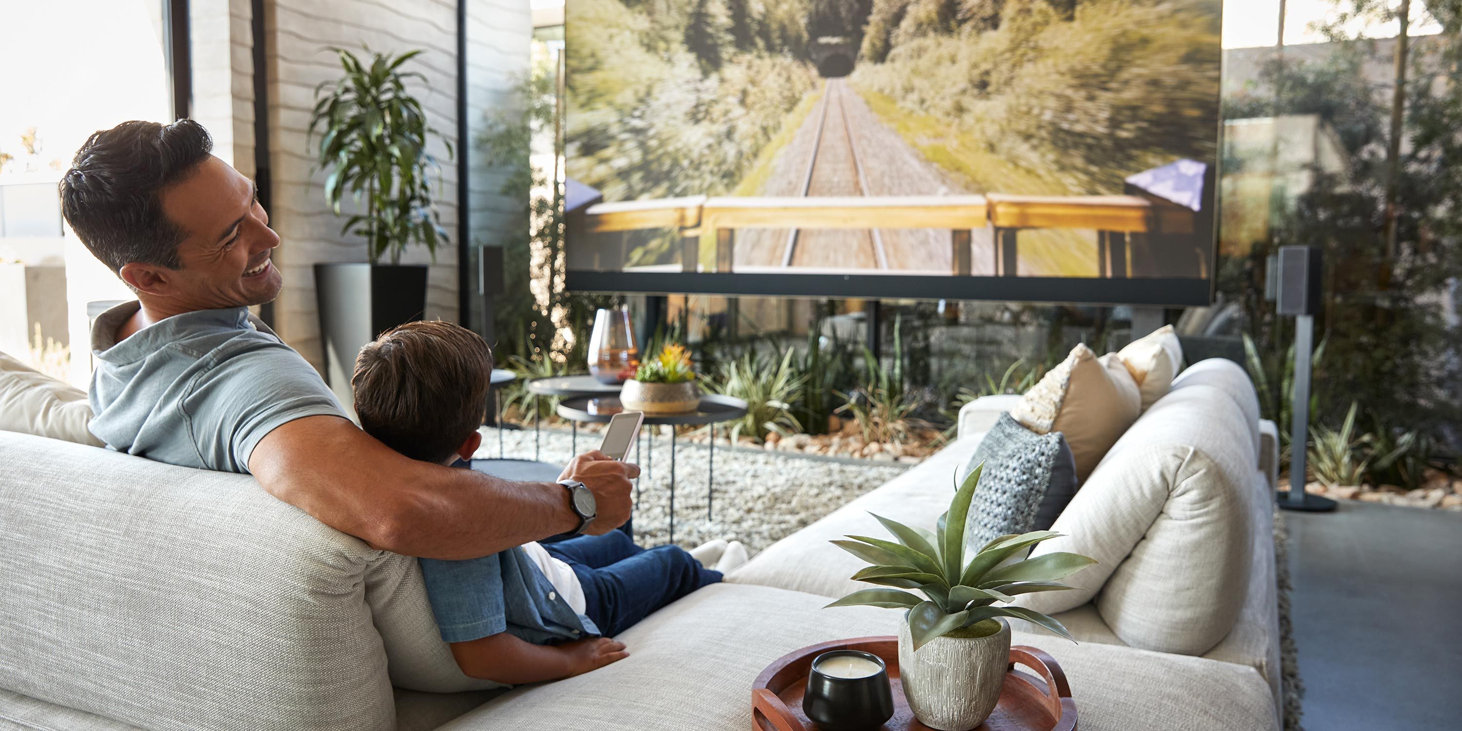 Father and son watching TV in a spacious and bright family room with lush indoor plants.